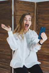 Emotional young woman in a white cotton shirt gesticulates and looks at the screen of a modern...
