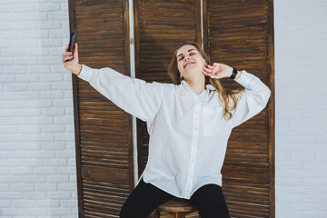 Emotional young woman in a white cotton shirt gesticulates and looks at the screen of a modern...