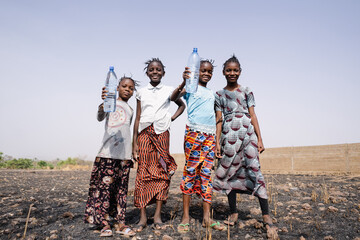 Four beautiful black African girls in casual wear standing in a dry field proudly holding up water...
