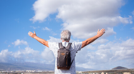 Back view of senior caucasian man with backpack on shoulders walking outdoors in countryside with open arms. White-haired old man enjoying freedom travel and nature. Copy space