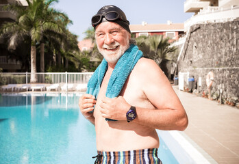 Smiling senior man on the edge of the swimming pool, wearing goggles and swimming cap holding a...