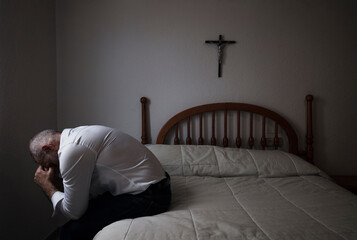 Adult man in white shirt in room sitting on bed with cross on wall