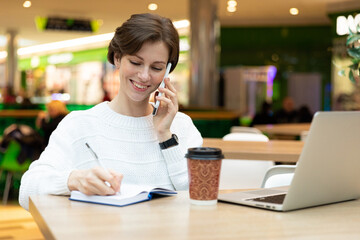 Young attractive positive smiling brunette woman sitting at a shopping center at a table with coffee paper cup and working at a computer laptop, using mobile phone. Freelance and business concept