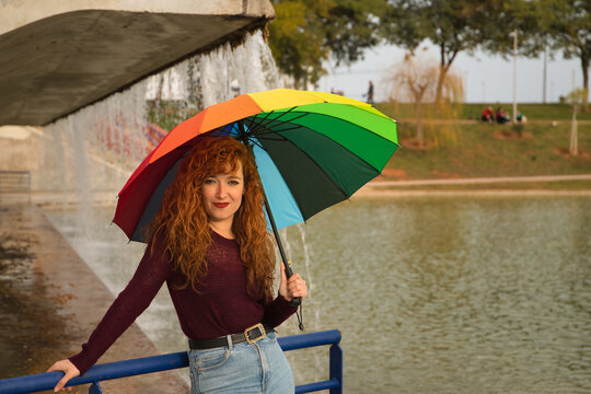 Portrait of young woman, red hair, freckles, with a rainbow umbrella, happy, in an outdoor park. Concept color, happiness, well-being, fun, rain.