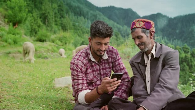 smiling Indian elderly male shepard with his young son sitting together using a Mobile phone while grazing sheeps in rural area of remote greenery mountainous region. 