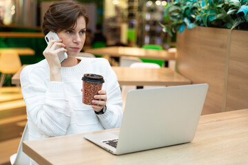 Young beautiful woman sitting at a shopping center at a table and working at a computer laptop, using mobile phone. Freelance and business concept