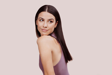 Close up shot of young smiling woman posing with head on shoulder looking away wears purple sleeveless top. Healthy skin, beauty portrait, smiling female with cosmetic at studio over beige background.