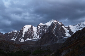 Obraz premium Dark atmospheric alpine landscape with snow-covered mountain top under night sky. Awesome scenery with beautiful pointy peak with snow and high snowy mountain wall with dark low clouds.