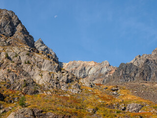 Bright mountain landscape with sharp rocks above yellow slope under blue sky with moon in sunny day. Colorful scenery with gold sunlit sharp rocky mountains. High rocky mountains in golden sunlight.