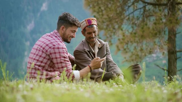 Indian rural father son duo sitting on a meadow smiling watching a funny or comedy video on mobile phone using OTT platform or social media  spending quality time together in a mountainous region