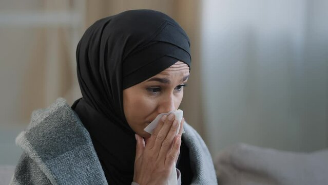 Close up sick upset young girl in hijab sneeze wipe nose with paper napkin sad unhealthy muslim woman drinking hot tea suffering from seasonal cold feel virus flu symptoms coronavirus epidemic concept