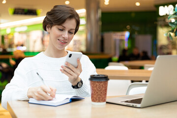 Fototapeta na wymiar Young attractive brunette woman sitting at a shopping center at a table with coffee paper cup and working at a computer laptop, using mobile phone. Freelance and business concept