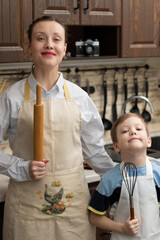A young beautiful mother with her son in aprons with a rolling pin and a whisk in their hands at home in the kitchen prepare food against the background of kitchen utensils. Selective focus. Portrait