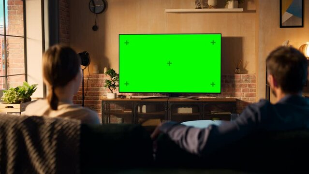 Authentic Couple Spending Time at Home, Sitting on a Couch and Watching TV with Green Screen Mock Up Display in Their Stylish Loft Apartment. Man and Woman Streaming Movie or Show. Shot From Back.