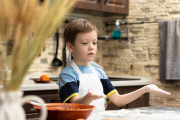 A cute boy of 7 years old in an apron sprinkles flour on the table at home in the kitchen against the background of kitchen utensils. Selective focus. Portrait