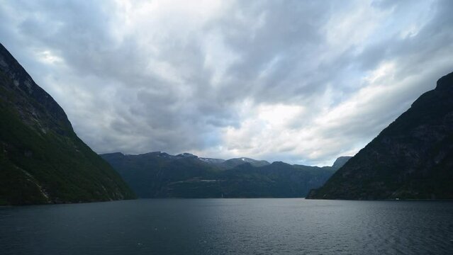 Cinematic time-lapse footage where the camera is placed in the front of a cruise ship, that is leaving the most famous fjord of Norway, Geirangerfjord. The sky is mostly covered with clouds.