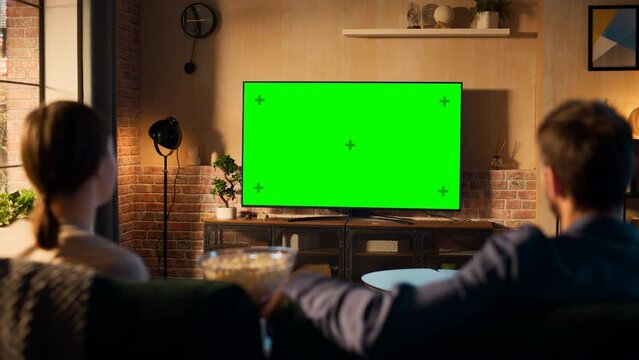 Young Couple Spending Time at Home, Sitting on a Couch and Watching TV with Green Screen Mock Up Display in Their Stylish Loft Apartment. Man and Woman Streaming Movie or Show. Shot From Back.