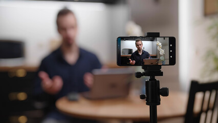 Businessman with laptop recording video with smartphone in kitchen at home