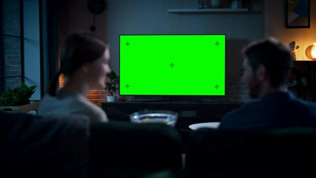 Authentic Couple Spending Time at Home, Sitting on a Couch and Watching TV with Green Screen Mock Up Display in Their Stylish Loft Apartment. Man and Woman Streaming Movie or Show. Shot From Back.