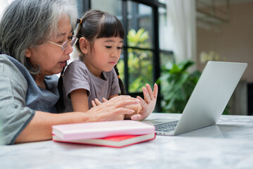 Asian Grandmother with her two grandchildren having fun and playing education games online with a computer notebook at home in the living room. Concept of online education and caring from parents.