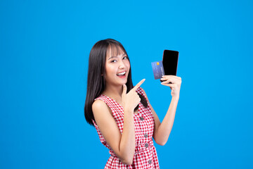 Portrait Asian happy young girl smiling cheerful and showing plastic credit card while holding mobile phone isolated on blue background