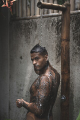 Obraz premium Shirtless muscular latin american male with tattoos standing taking shower outdoors after workout on the tropical leaves background. 