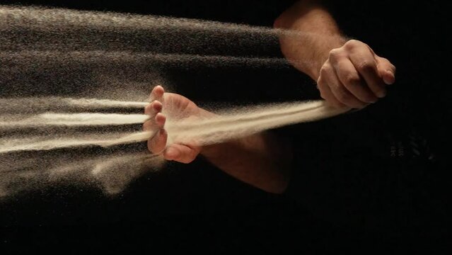 Stream of dry sand pours into palm of man and is spilling through his fingers on black background. Small grains of sand are sifted and scatter in different directions in slow motion. Close up.