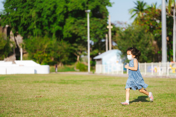 Fototapeta premium Portrait cute kid girl running on green grass. Child wearing face mask prevent virus and air pollution PM2.5. Children playing sport in summer time. Hot weather. Empty space for enter text.