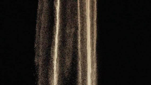Particles of white dry sand are pouring down on black studio background. Close up shot stream of flowing natural sand grains. The sand flows down. Slow motion.