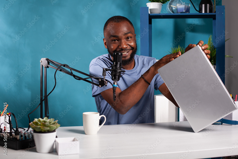 Poster smiling influencer recording silver laptop giveaway for website sitting at desk in vlogging studio w