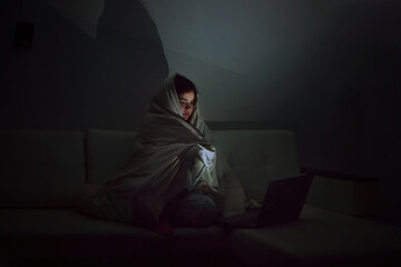 Selective focus on a woman who sits in the dark in a dark room on the couch, covered with a...