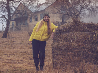 A smiling woman stands leaning on a haystack in an abandoned area against the background of an old...
