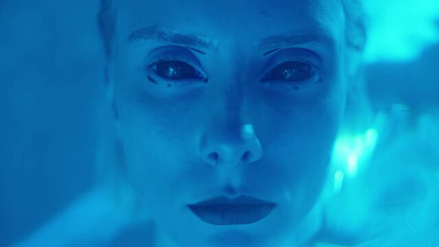 Close up shot of young woman with blackout contact lenses and alien makeup looking at camera while posing in dark studio filled with laser light