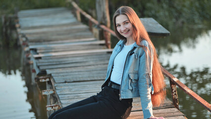 A young girl with long hair poses by the bridge on the river.