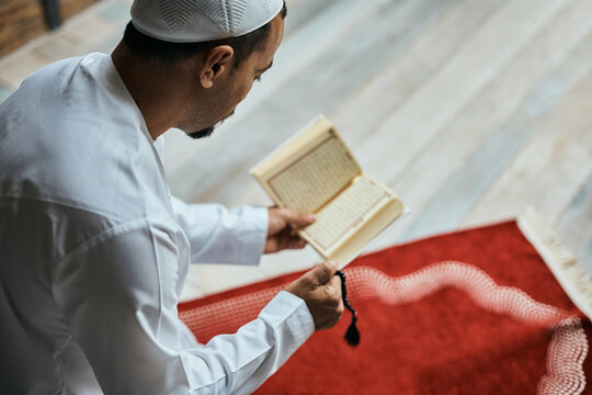 Religious Muslim man reads from Koran while holding prayer beads at home.