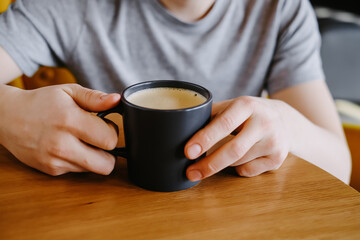 Hands of a man in a grey t-shirt, holding a cup of coffee on a wooden table