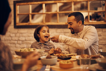 Happy Middle eastern father feeds his daughter during dinner at dining table on Ramadan.
