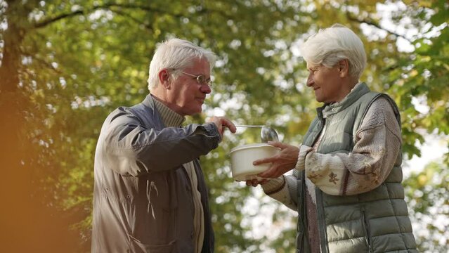 European elderly grey-haired man helps poor homeless people by volunteering and pouring nutritious soup into the plastic bowl that belongs to an elderly woman in need. High quality 4k footage
