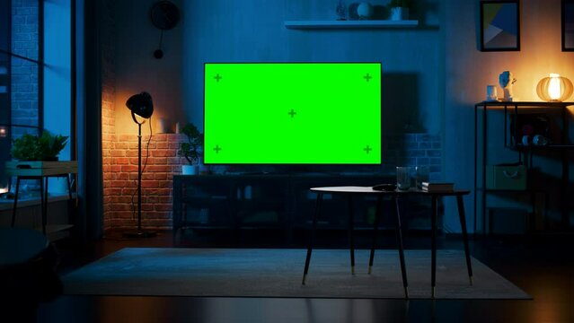 Stylish Loft Apartment Interior with TV Set with Green Screen Mock Up Display Standing on Television Stand. Empty Living Room at Home with Chroma Key Placeholder on Monitor. Still Night Shot.