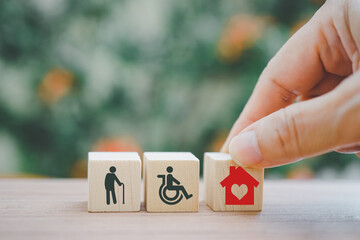 hand holding red house with heart  on row of wood cubes , elderly and disability person icon on blurred nature outdoor background ,home care for aging people concept