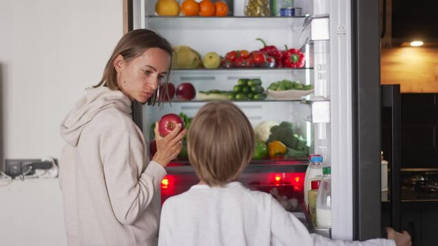 Boy opening refrigerator door while standing in kitchen. Refrigerator full of fruits and vegetables. Healthy lifestyle concept.