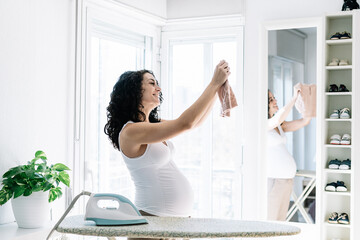 young pregnant woman ironing a small sweater of her future baby, preparing and organizing the room of the newborn