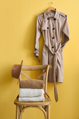 Wooden chair with sweaters, hat and trenchcoat hanging on yellow background