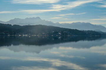 Obraz premium View on Woerthersee in Poertschach in Carinthia, Austria.The calm surface of the lake is reflecting the mountains, sunbeams and clouds. Clear and sunny day. Karawanks mountain range. Alps, Lake Woerth