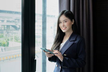 Fototapeta na wymiar Young businesswoman using smartphone working in hotel room during her business travel. Businesswoman working from hotel room on business trip standing by the window in sunny day.
