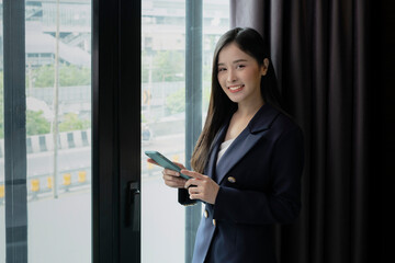 Young businesswoman using smartphone working in hotel room during her business travel. Businesswoman working from hotel room on business trip standing by the window in sunny day.