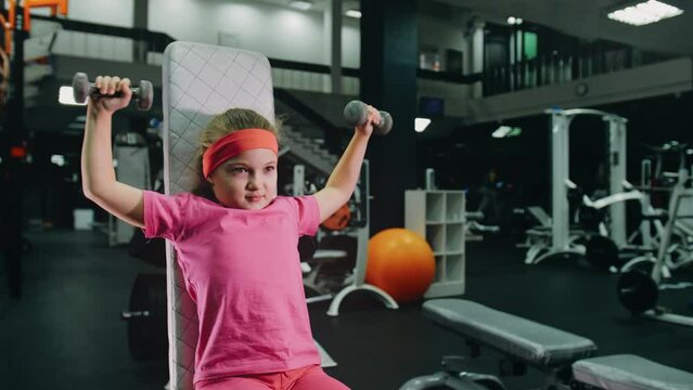 Fit Athletic Baby Girl Work out with Dumbbell, Doing Her Fitness Exercise. Children Activity Training in Modern Gym. Sports Child Workout in Exercising with Barbell, Fitness Center.