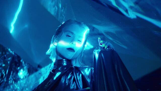 Stylish alien woman with dark lipstick and blackout contact lenses wearing metallic gown posing for camera in studio with liquid sky laser light