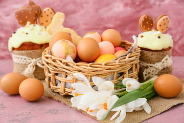 Gift basket with painted Easter eggs, flowers and cakes on pink background