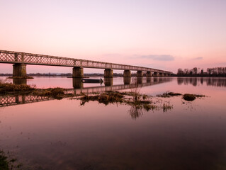 Pont en treillis enjambant un fleuve aux eaux calmes à la lumière du soir et avec les reflets mauve magenta à la surface. Grand Pont de Mauves sur Loire
